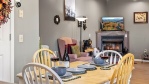 A dining area with a wooden table set for four, surrounded by white chairs. A fireplace with a painting above it and a television on a stand are visible in the background.