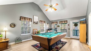 A pool table in a room with wooden floors, a ceiling fan, and plaid curtains on the windows.