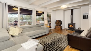 Un salon chaleureux avec un canapé gris, un divan en cuir noir et une cheminée. De grandes fenêtres offrent une vue sur l’extérieur.