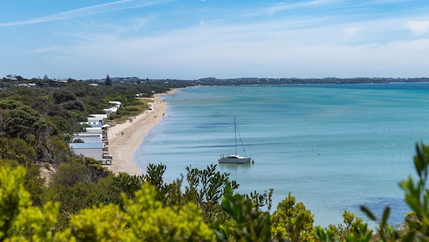 Une vue côtière avec une plage, un voilier et une rangée de bâtiments le long de la côte.
