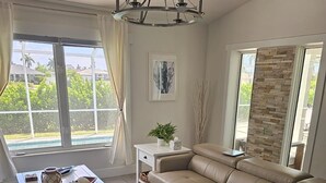 A living room with a beige sofa, a white coffee table, and a chandelier. There is a large window with curtains, a stone wall, and a painting on the wall. - Beautiful house and great neighborhood! (Marco Island)