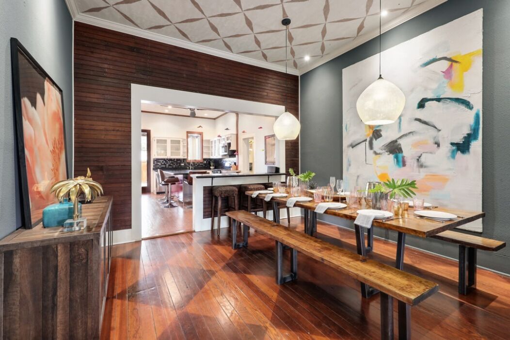 Een moderne eethoek met houten tafels en banken, een keuken op de achtergrond, en een groot abstract schilderij aan de muur.
