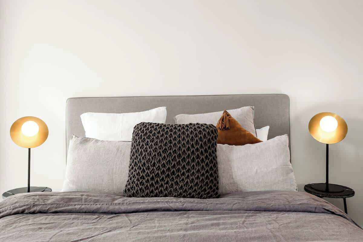 A neatly made bed with a grey headboard, two bedside lamps, and a variety of pillows.
