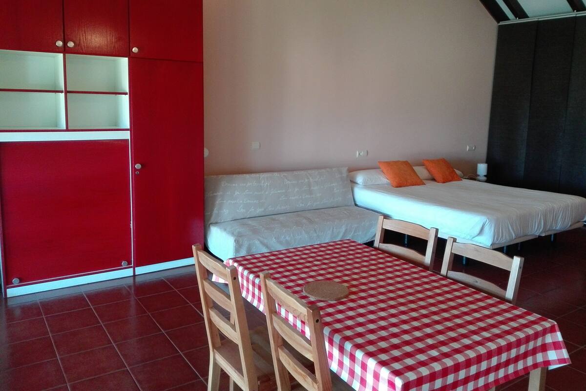 A room with a red wardrobe, a bed with orange pillows, a checkered table, and a wooden chair.
