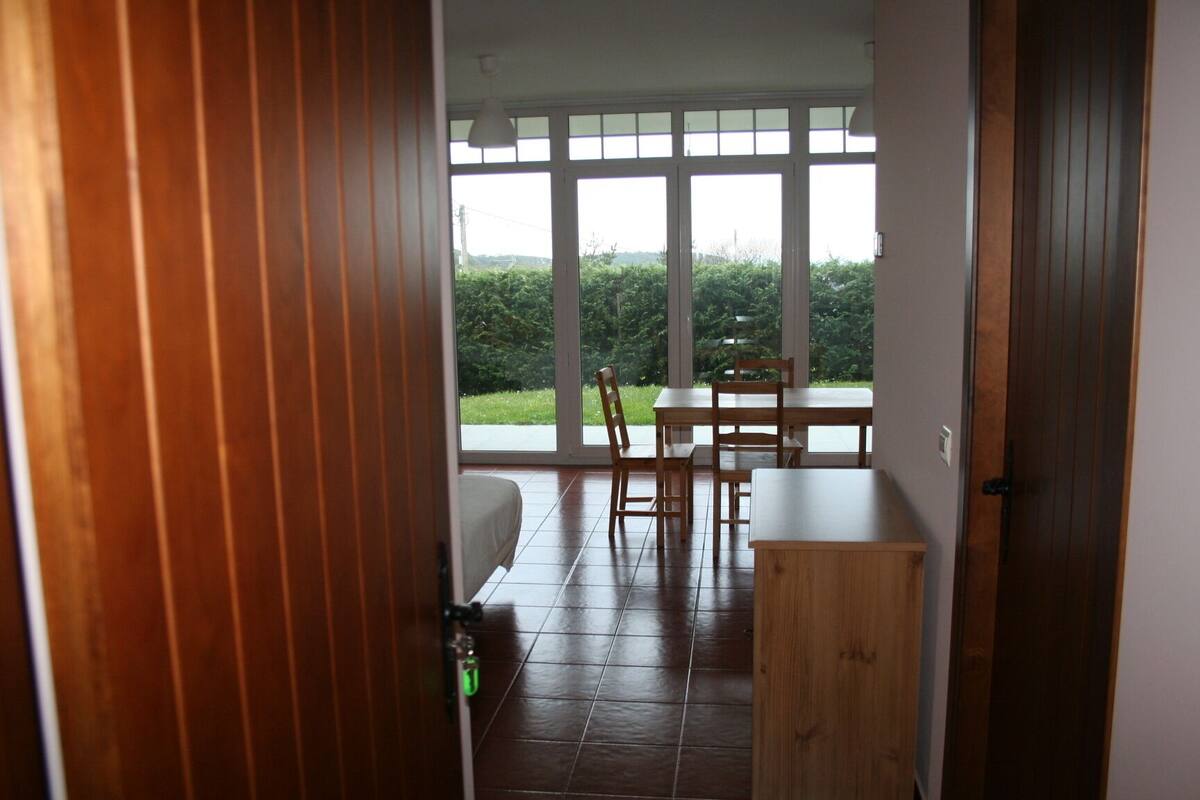 A room with a wooden door, a small table, and a chair. There is a window with a view of greenery outside.