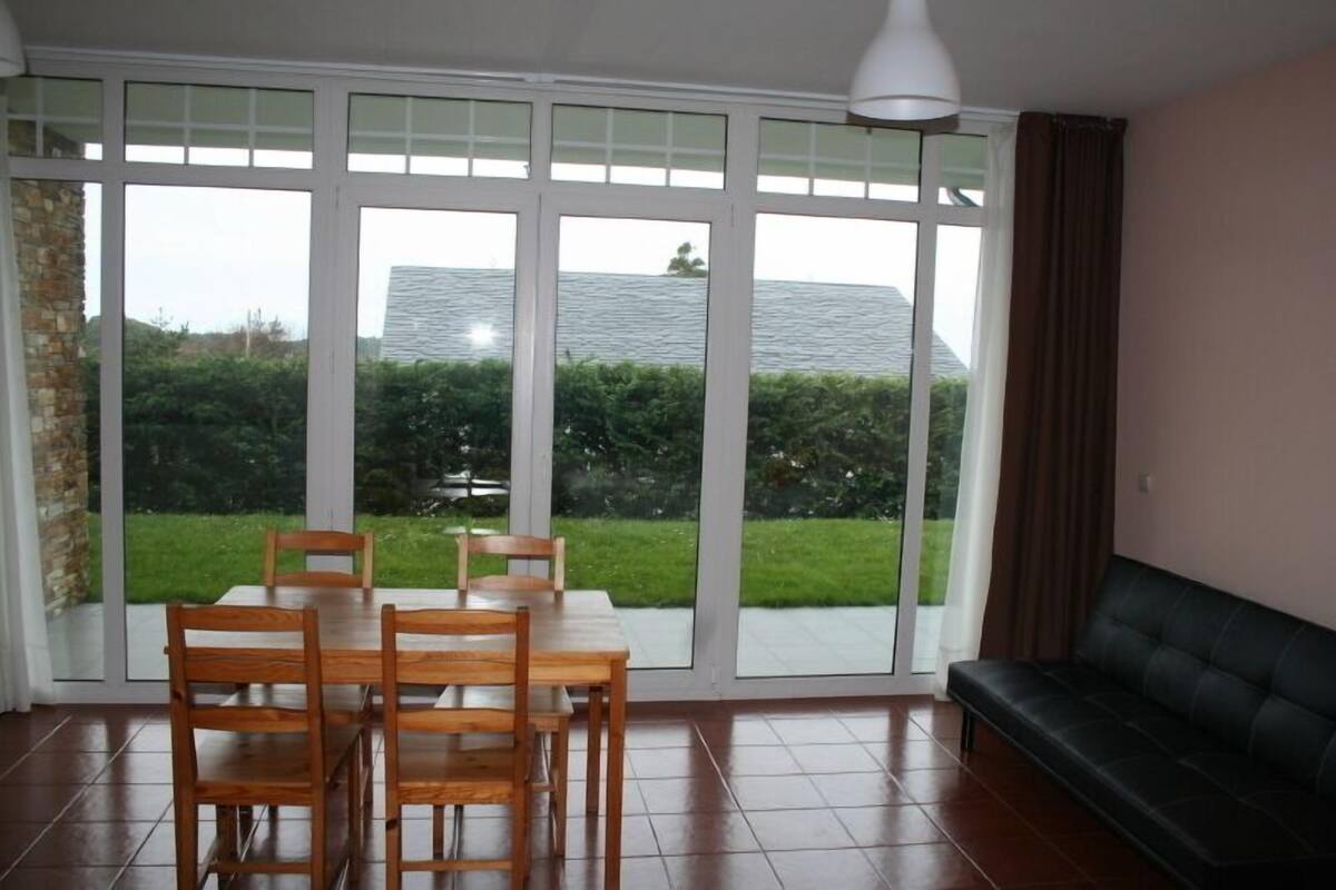 A room with a dining table and chairs, a black sofa, and large windows with a view of a green landscape and a building.