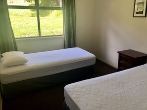 Una habitación con dos camas, una mesita de noche de madera, un ventanal con cortinas y un cuadro en la pared.
