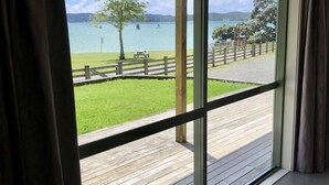 A room with a large sliding glass door leading to a deck with a wooden railing, a picnic table, and a grassy area with a fence. - Relax Beachside with your Family and Friends (Oakura)