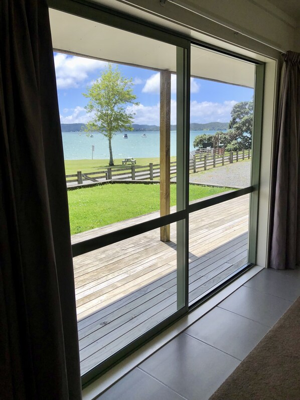 A room with a large sliding glass door leading to a deck with a wooden railing, a picnic table, and a grassy area with a fence.