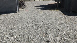 A gravel path leading to a beach area with a small building, a tree, and a boat in the background. - Relax Beachside with your Family and Friends (Oakura)