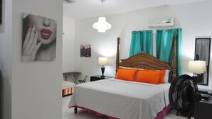 Ein Schlafzimmer mit einem Bett, zwei Nachttischen, einem Schreibtisch, einem Stuhl, einem Standventilator, einer Deckenleuchte und einer Wand-Klimaanlage.