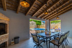 A dining area with a brick fireplace, a table with chairs, and a view of an outdoor swimming pool. - LUXURY VILLA ON SPACIOUS PRIVATE PROPERTY  (Rakalj)