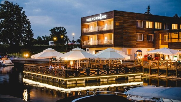 A hotel with a wooden facade, outdoor seating area with umbrellas, and a dock with boats. - Hotel Port 110 (Ilawa)