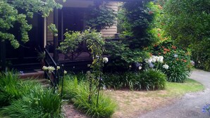 A house with a porch surrounded by lush greenery and flowering plants. - ROSE AT LEAFIELD COTTAGES (Dixons Creek)