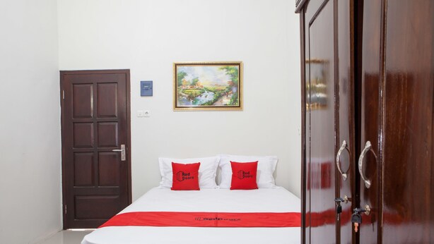 A hotel room with a bed, two red pillows, a painting on the wall, and a wooden wardrobe. - RedDoorz near Banyuwangi Baru Station (Banyuwangi)