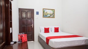 A hotel room with a wooden door, a bed with white bedding and red accents, a framed painting on the wall, and a red bag on the floor. - RedDoorz near Banyuwangi Baru Station (Banyuwangi)