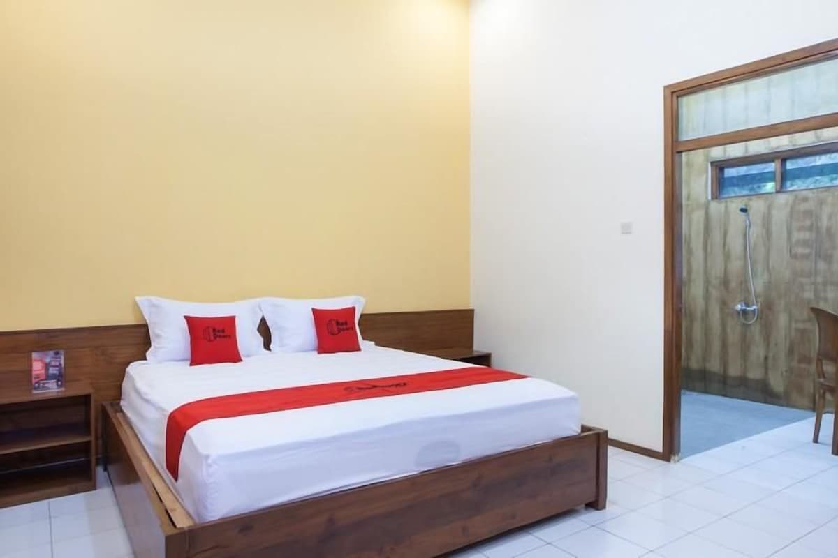 Una cama individual con ropa blanca y detalles rojos, cabecera de madera y una manta roja.