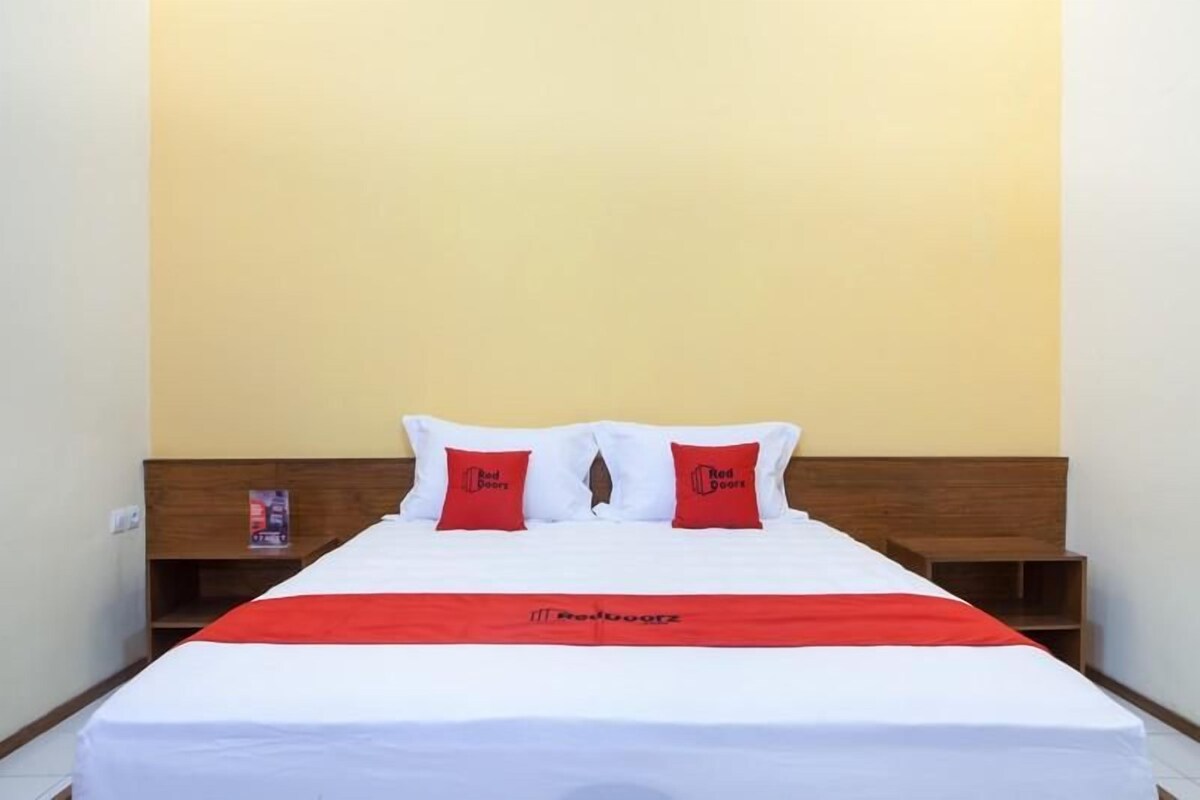 Une chambre d’hôtel avec un lit, deux oreillers rouges, une tête de lit en bois, une couvre-lit rouge et une table de chevet avec un livre.