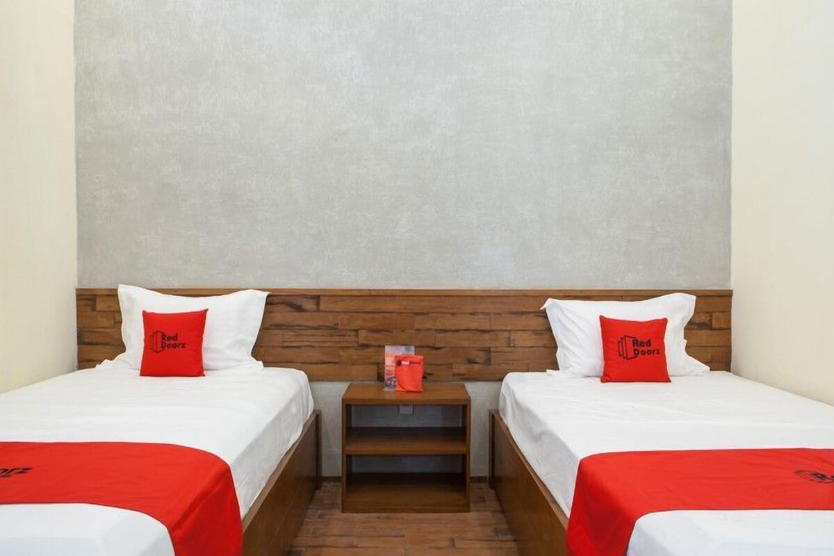 Une chambre d’hôtel avec deux lits, chacun recouvert d’une courtepointe blanche et agrémenté d’un oreiller rouge. On y trouve une tête de lit en bois, une petite table de chevet en bois avec un sac rouge, et un tableau encadré posé sur la table de chevet.