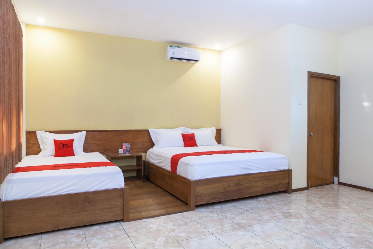 Une chambre d’hôtel avec deux lits, une tête de lit en bois, une table de chevet, un plaid rouge et un appareil de climatisation.