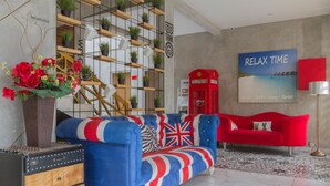 Une pièce avec un canapé bleu et rouge, une cabine téléphonique rouge, un mur orné d’une affiche « MOMENT DE DÉTENTE » et un mur végétal.