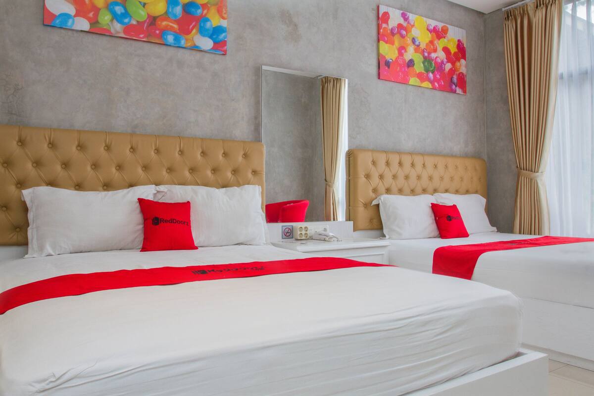 Habitación de hotel con dos camas, cada una con cabecero capitonado, ropa de cama blanca y detalles en rojo. Hay cuadros coloridos en las paredes y un espejo grande.