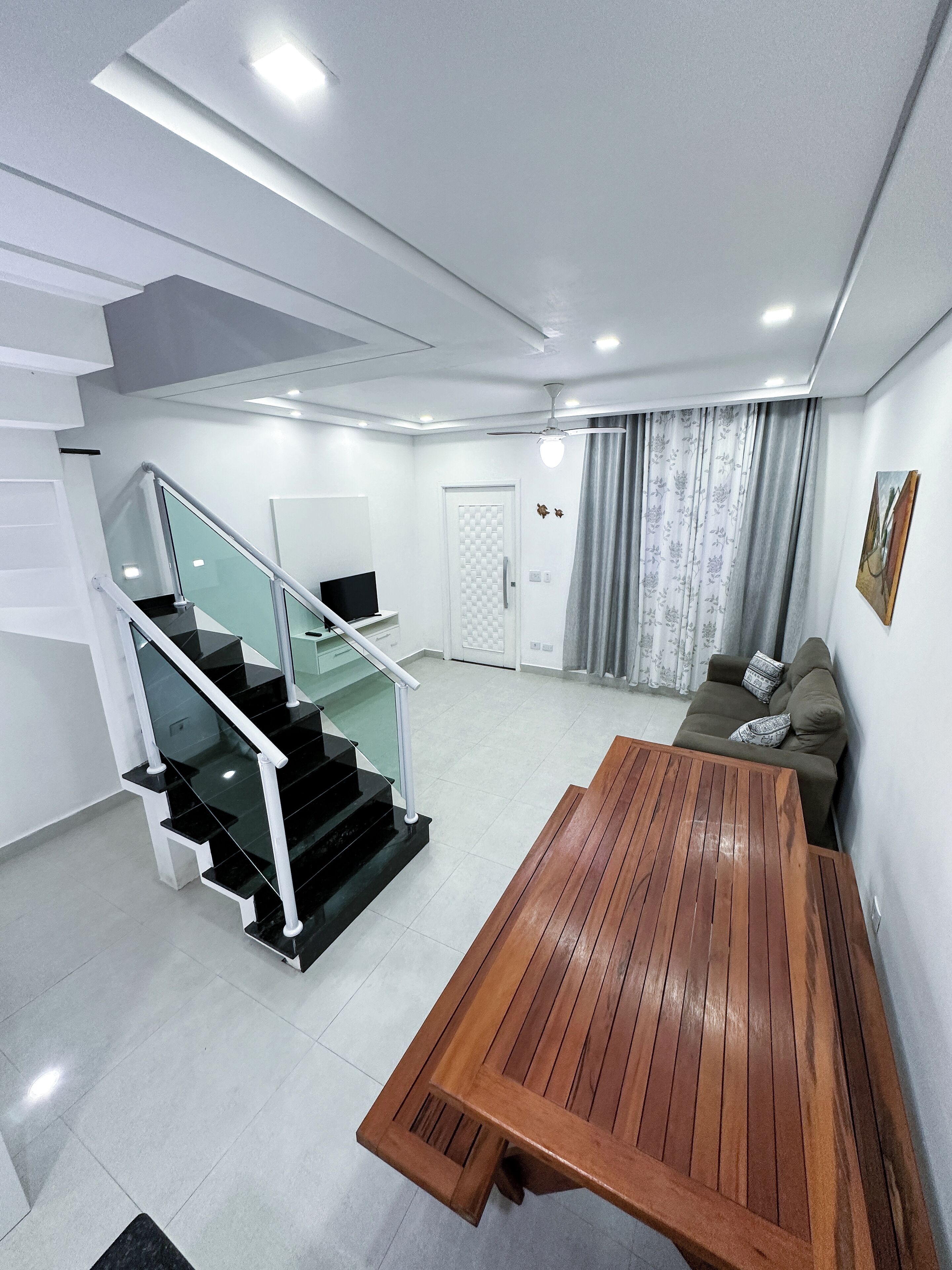A modern living room with a wooden table, a sofa, a staircase with glass railings, and a door.