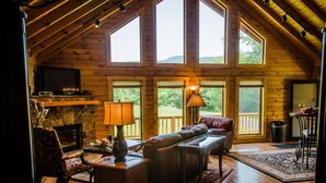 A cozy cabin interior with a fireplace, leather sofa, and large windows offering a view of the outdoors. - ✧Royale Cabin- KING bed, hot tub, NO CLEANING FEE (Dryfork)