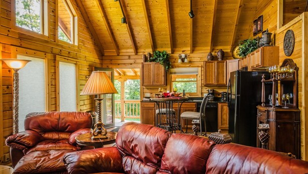 A cozy log cabin interior with a leather sofa, wooden ceiling beams, and a kitchen area. - ✧Royale Cabin- KING bed, hot tub, NO CLEANING FEE (Dryfork)