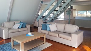 A modern living room with two beige sofas, a wooden coffee table, and a blue rug. A staircase with blue railings is visible in the background.