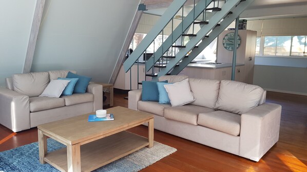 A modern living room with two beige sofas, a wooden coffee table, and a blue rug. A staircase with blue railings is visible in the background.