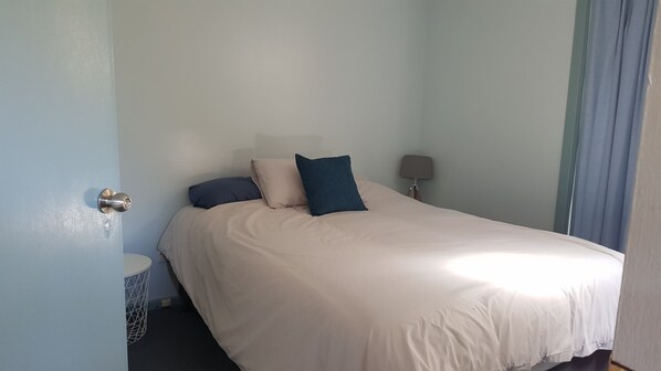 A neatly made bed with a white duvet and two pillows, a bedside table with a lamp, and a window with curtains.