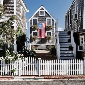 A two-story house with a front porch, a white picket fence, and an American flag.