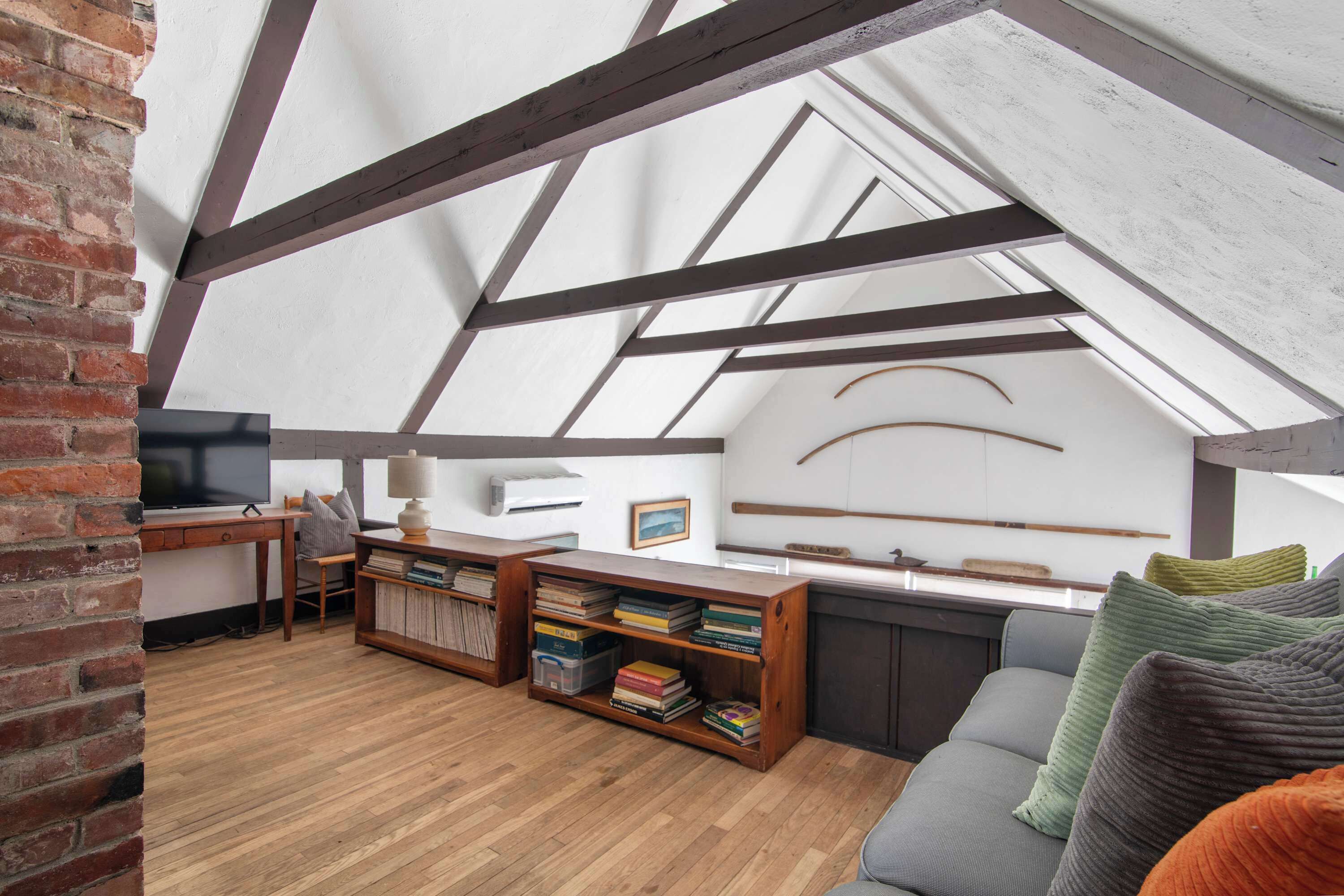 Una habitación estilo loft con vigas de madera a la vista, pared de ladrillo, un escritorio con computadora, una estantería y un sofá con cojines.