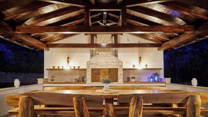 Un espace de restauration extérieur rustique, doté d’une cheminée en pierre, de bancs en bois et d’un plafond haut avec des poutres apparentes.