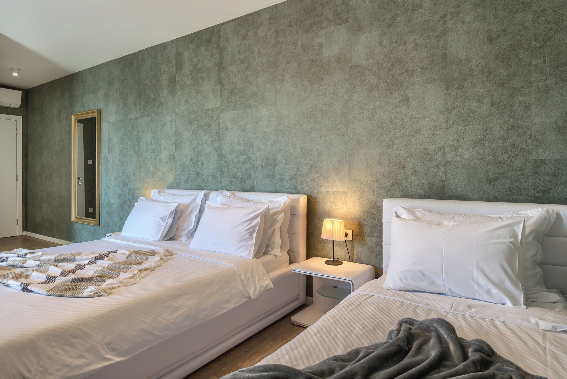 Une chambre d’hôtel avec deux lits, un grand miroir, une table de chevet avec une lampe et un mur vert à texture.