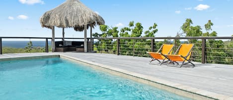 A swimming pool with two orange lounge chairs and a thatched-roof structure.