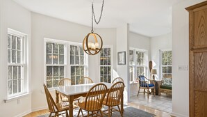 Uma sala de jantar com uma mesa branca e cadeiras de madeira, janelas amplas e um lustre.