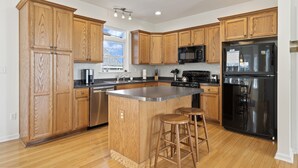 Uma cozinha moderna com armários de madeira, uma ilha central com banquetas, eletrodomésticos de aço inoxidável e uma janela que proporciona iluminação natural.