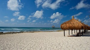 A beach with thatched umbrellas and lounge chairs, a clear blue sky, and a distant pier.