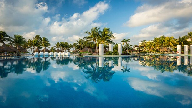 A large, clear swimming pool surrounded by palm trees and lounge chairs, reflecting the sky and clouds.