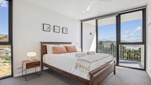 A bedroom with a large bed, a nightstand, a lamp, and a view of a balcony with a scenic landscape. - Chilam B embraces open plan living with a combined kitchen/ dining/ living area (Jindabyne)