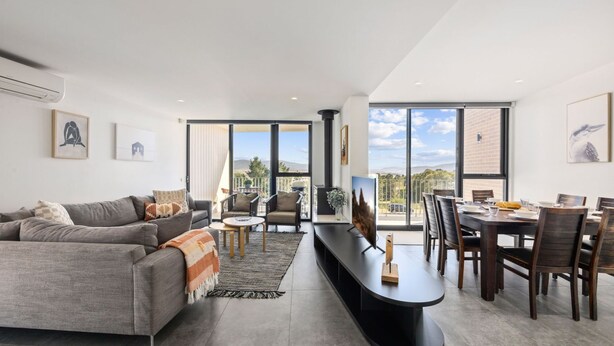 A modern living room with a grey sofa, a coffee table, and a dining area with wooden chairs and a table. - Chilam B embraces open plan living with a combined kitchen/ dining/ living area (Jindabyne)