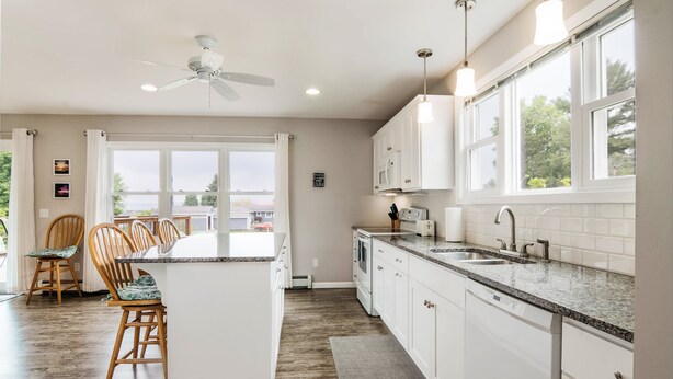 A modern kitchen with white cabinets, granite countertops, and a central island. There is a ceiling fan, pendant lights, and a view of the outdoors through large windows. - Beautiful Cottage Overlooking Bear Lake (Bear Lake)