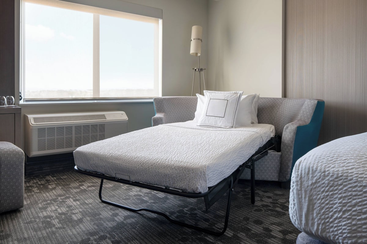 A hotel room with a bed, two armchairs, a floor lamp, and a large window.