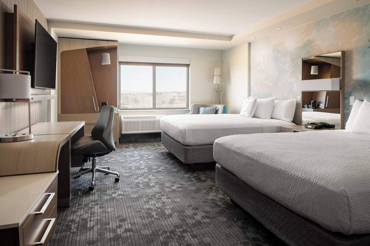 A hotel room with a desk, two beds, a TV, and a large window.