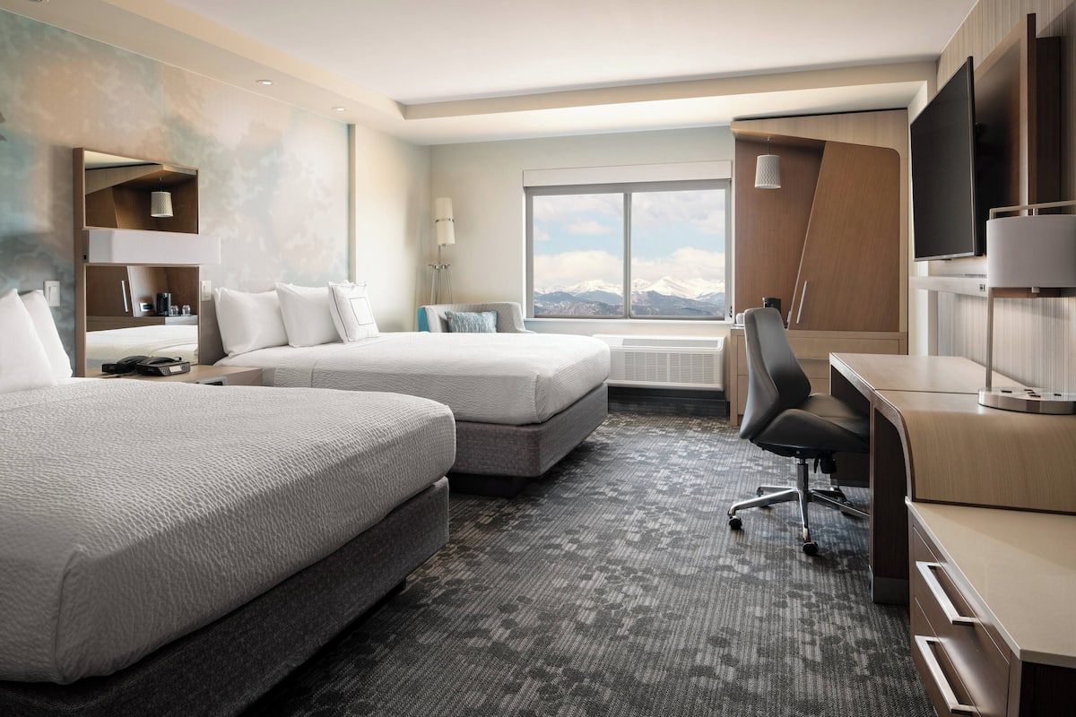 A hotel room with two beds, a desk, a chair, and a large window with a mountain view.