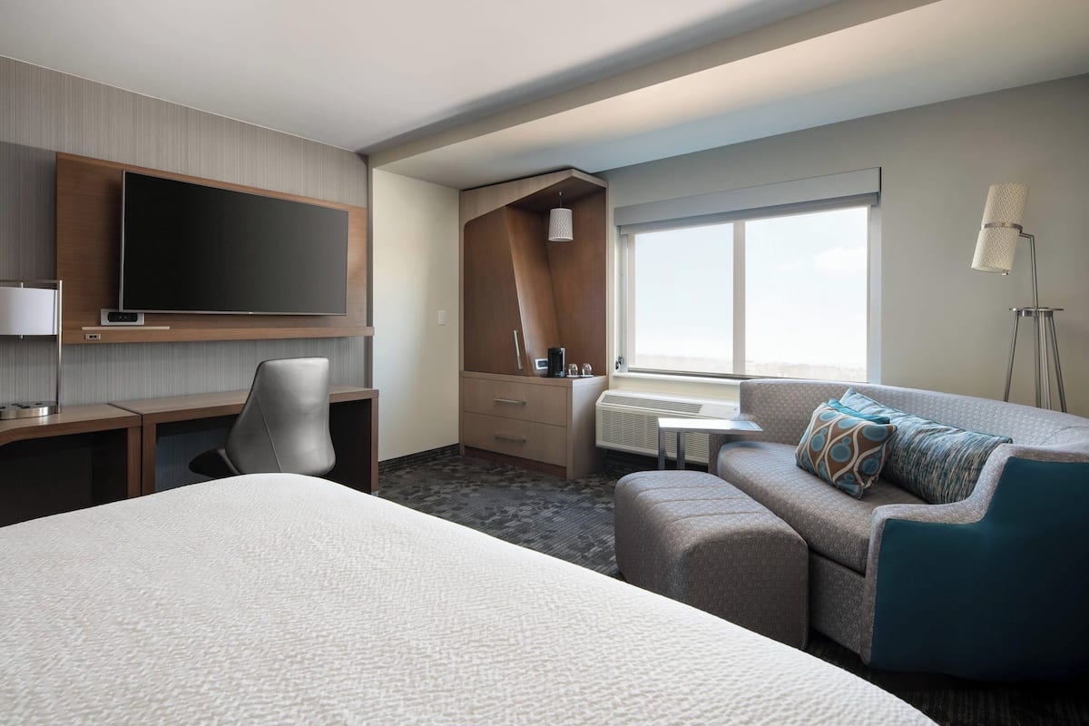 A hotel room with a large bed, a desk with a flat-screen TV, a sofa, a small table, and a window with a view.