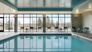 A swimming pool with clear blue water, surrounded by lounge chairs and tables, and large windows offering a view of trees and buildings. - Courtyard by Marriott Loveland Fort Collins (Loveland)