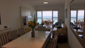 Un amplio salón con una larga mesa de comedor, asientos cómodos y ventanas grandes que ofrecen vista al mar.